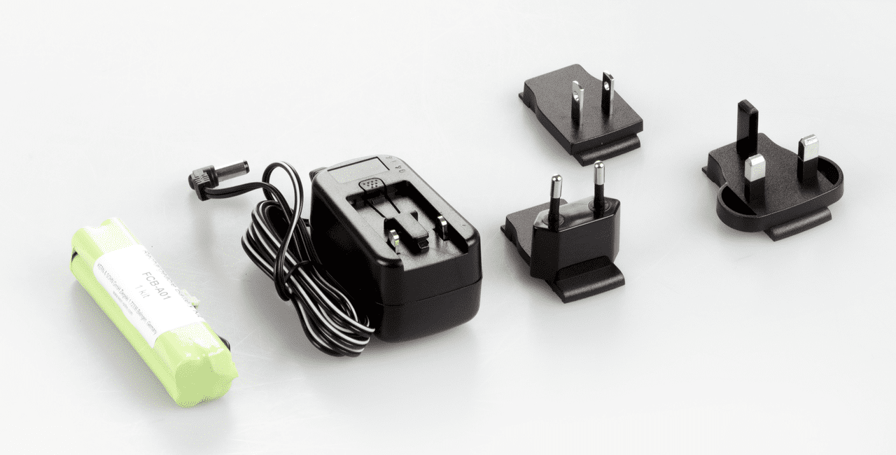 Akkubetrieb auf weißem Hintergrund. Das Set besteht aus (von Links nach Rechts im Bild): eine austauschbare Batterie, ein Hauptstecker mit aufgefaltetem Kabel, drei Adapter für EU, US und UK.