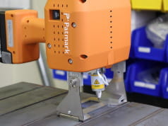 Oranger Abstandhalter im Einsatz auf einer Werkbank. Das Gerät zeigt nach unten, die Metallschiene steht dabei auf der Tischplatte.